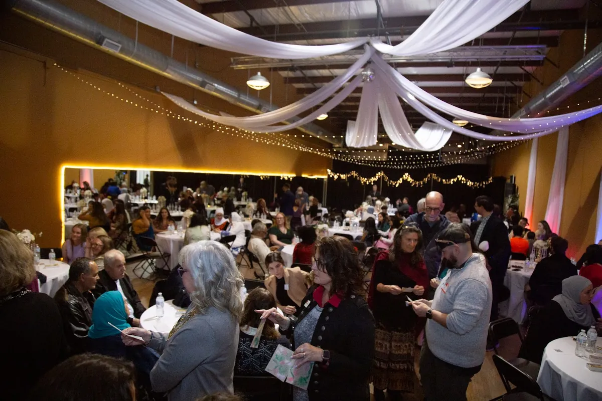 Guests talking together at an MMY Appreciation Iftar in a warmly lit hall with draped fabric and string lights