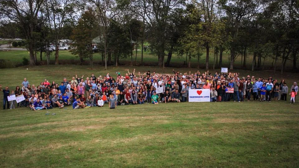 A large crowd of neighbors gathered on a grassy hillside holding a #MurfreesboroLoves banner with a heart