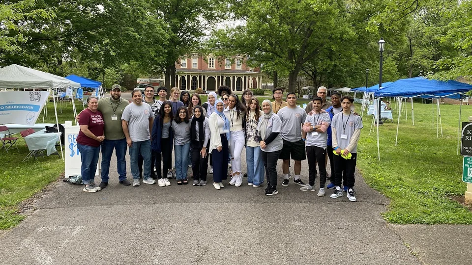 The NeuroDash setup team, about two dozen volunteers in gray shirts, standing together at Oakland Mansion with event tents and a NeuroDash banner behind them
