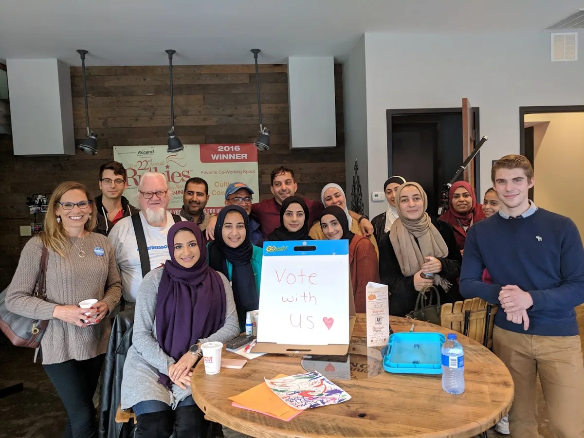 MMY members and community volunteers gathered around a 'Vote with Us' sign at a civic engagement event