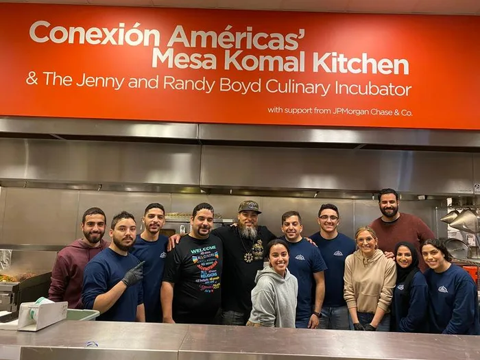 A group of volunteers smiling together in a commercial kitchen under a Conexión Américas Mesa Komal Kitchen sign